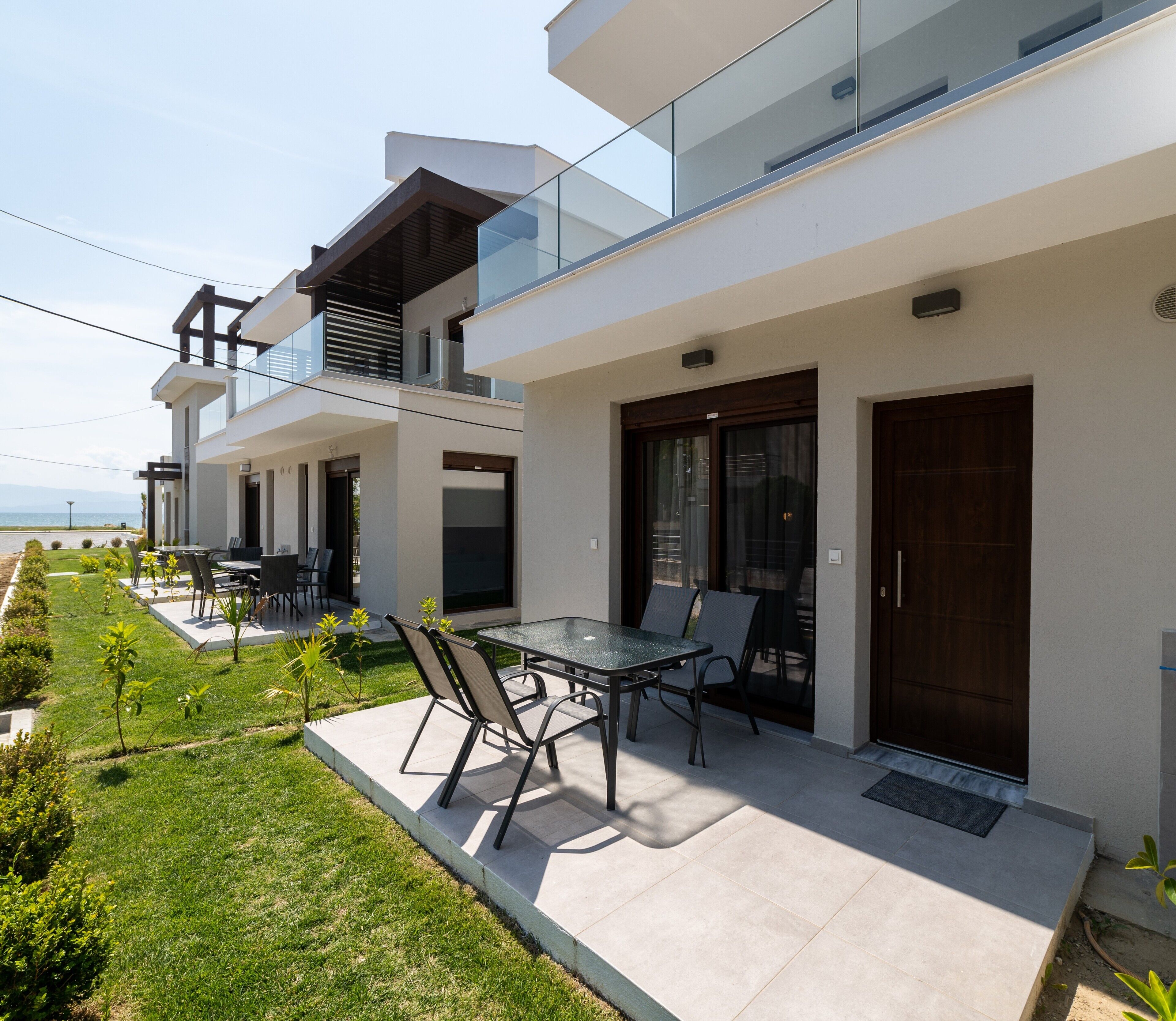 A modern two-story residential building with a balcony, a dining area with outdoor furniture, and a well-maintained lawn.