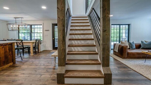 A spacious living area with a wooden staircase, a dining area, and a large window providing natural light. - Fall Brook: 3 bedroom cabin by waterfalls and hiking trail (Penn Yan)