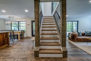 A spacious living area with a wooden staircase, a dining area, and a large window providing natural light.
