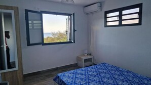A bedroom with a bed, a small bedside table, a window with a view, and an air conditioning unit.