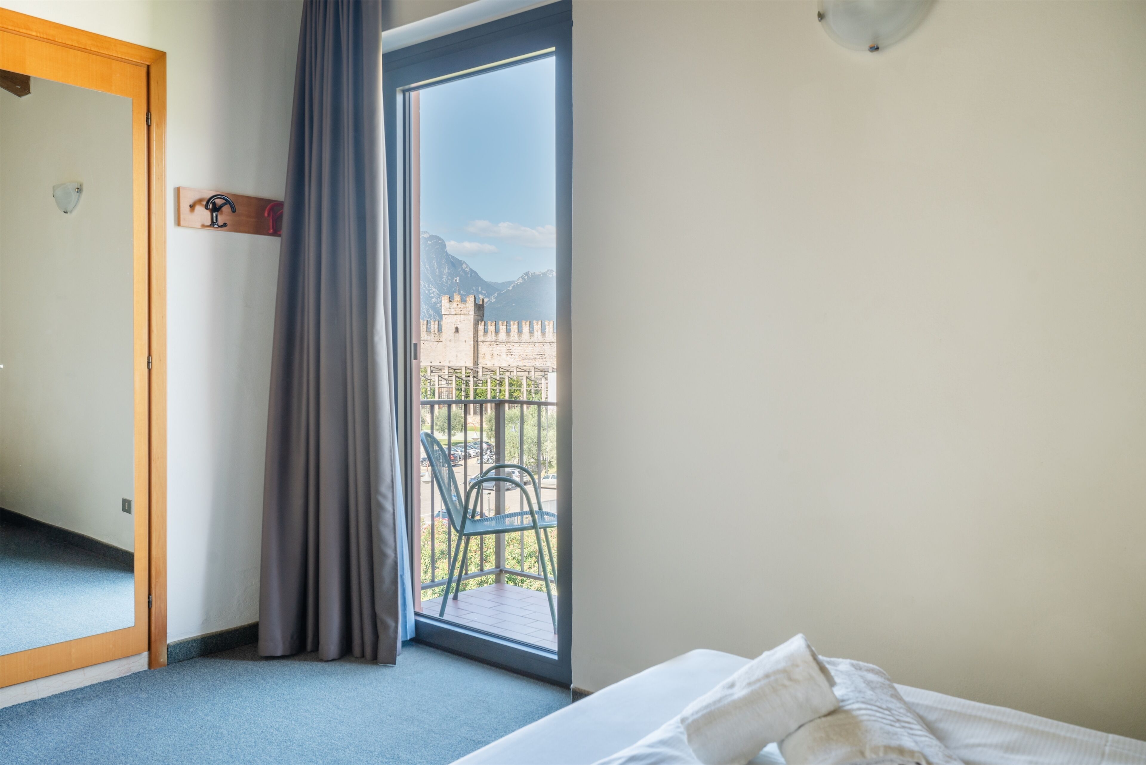 A hotel room with a bed, a window with a view of a castle, a balcony with chairs, and a coat rack.