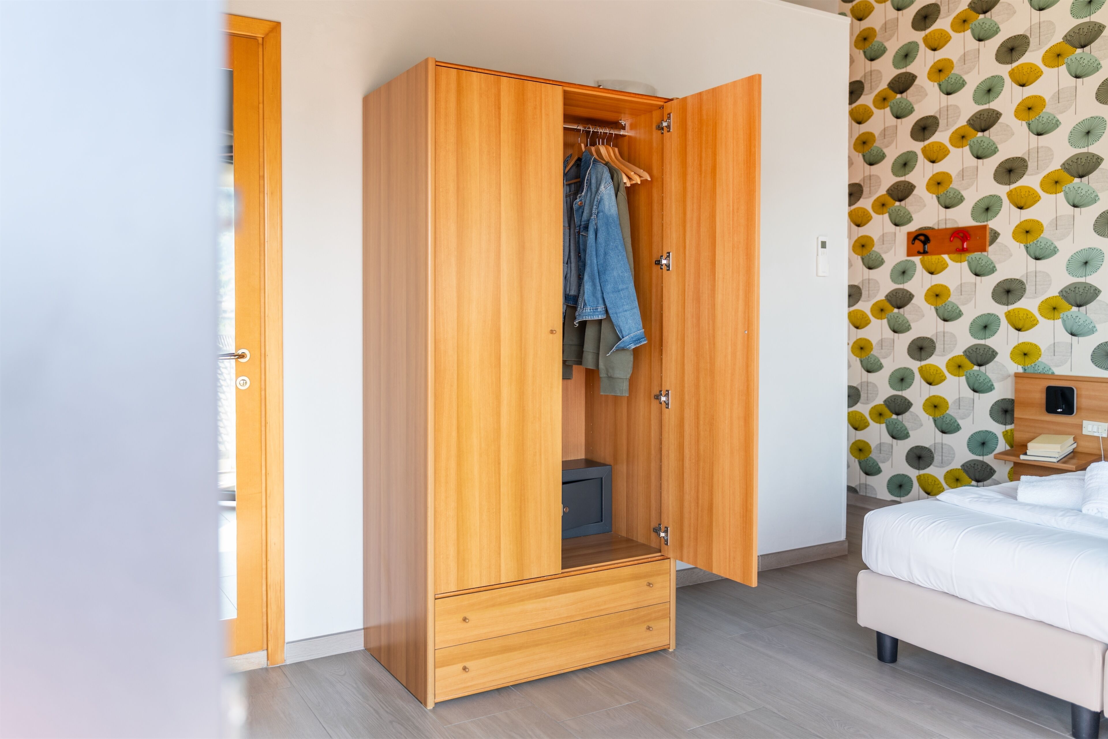 A bedroom with a wooden wardrobe, a bed, and a patterned wallpaper.