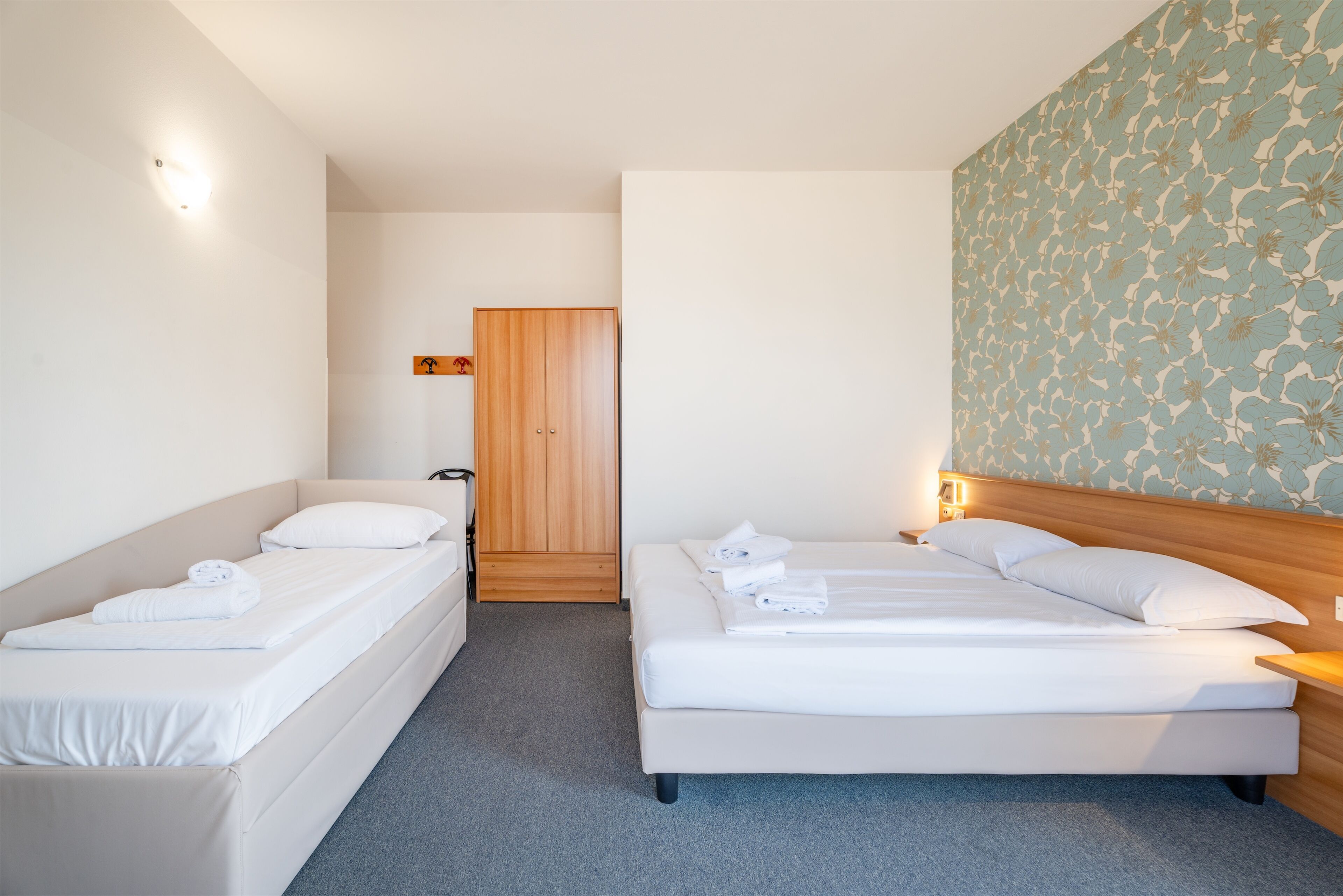 Ett hotellrum med två separata sängar, en garderob i trä, ett litet badrum och en blommig tapet på en av väggarna.