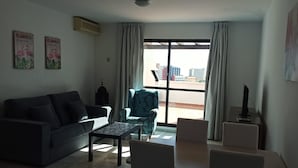 Un salon comprenant un canapé, un fauteuil à motifs, une petite table et un téléviseur posé sur un meuble. On y trouve également deux climatiseurs fixés au mur et une fenêtre avec des rideaux.