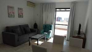 Un salon comprenant un canapé, des fauteuils, une table basse et une table à manger avec des chaises. Il y a une fenêtre avec des rideaux, un climatiseur fixé au mur et une lampe.