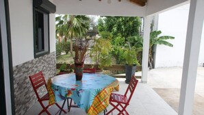 A covered patio with a round table and four red chairs, surrounded by potted plants and a view of a garden.