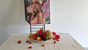 A table with a fruit and flower arrangement, a chair, and a painting on the wall.