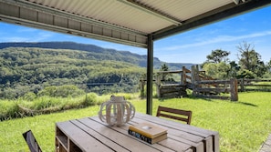 Relax in a serene outdoor setting with a wooden table and chairs under a covered area, surrounded by lush greenery and a scenic view.