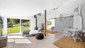 A modern bedroom with a large sliding glass door leading to a garden, a bed with a view, a rustic wooden table, and a ceiling fan. - Molla Eco Lodge Kangaroo Valley (Wattamolla)