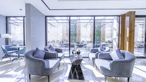 A modern hotel lobby with grey upholstered chairs, a black marble coffee table, and large floor-to-ceiling windows offering views of the outdoor terrace. - Mabaat - Al Malqa - 523 (Riyadh)
