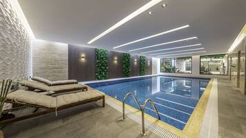 A modern indoor swimming pool with lounge chairs, a glass barrier, and a view of a gym area.