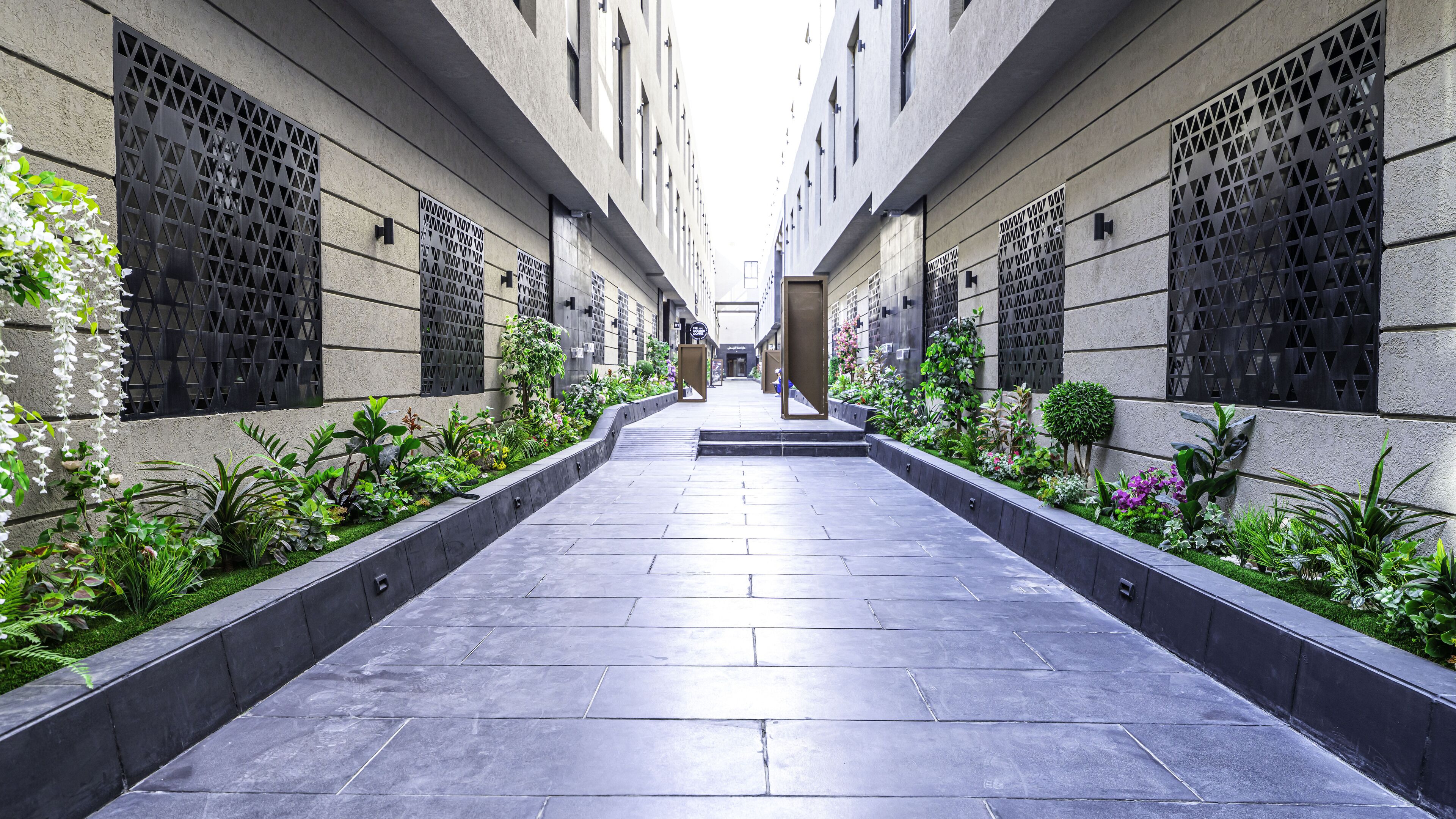 A narrow, paved walkway flanked by buildings with geometric window patterns and lush greenery.