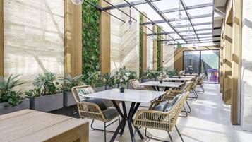 A modern outdoor dining area with wooden tables and wicker chairs, surrounded by greenery and a glass ceiling.