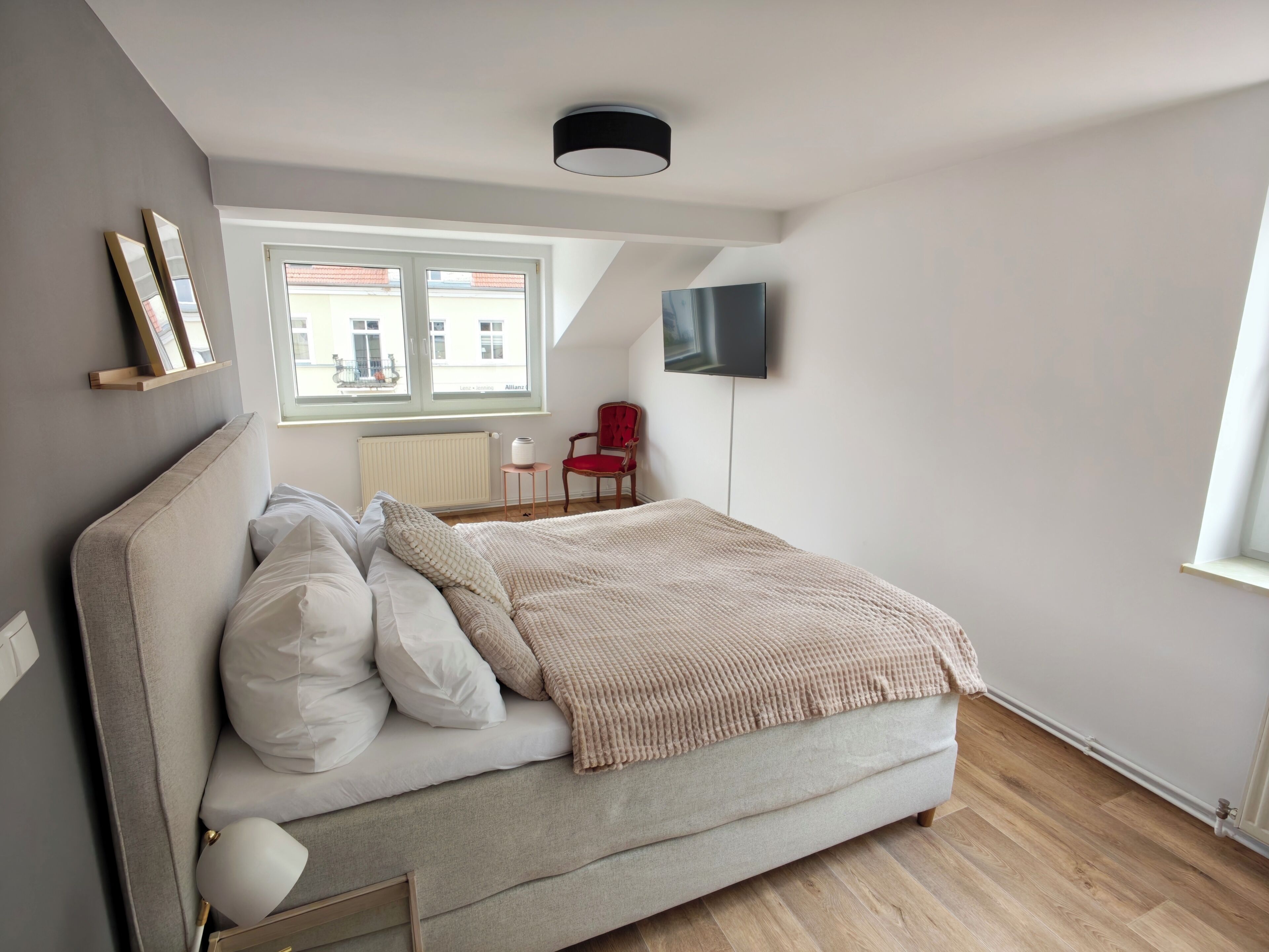 Un dormitorio con una cama, un televisor montado en la pared, una ventana y una silla.