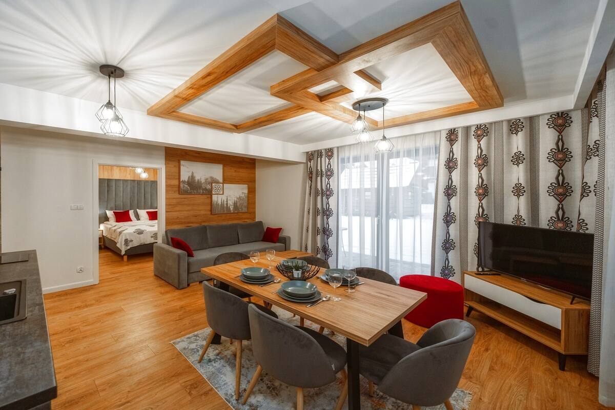 Uma sala de estar moderna com uma mesa de jantar de madeira, sofás cinzentos e uma televisão.