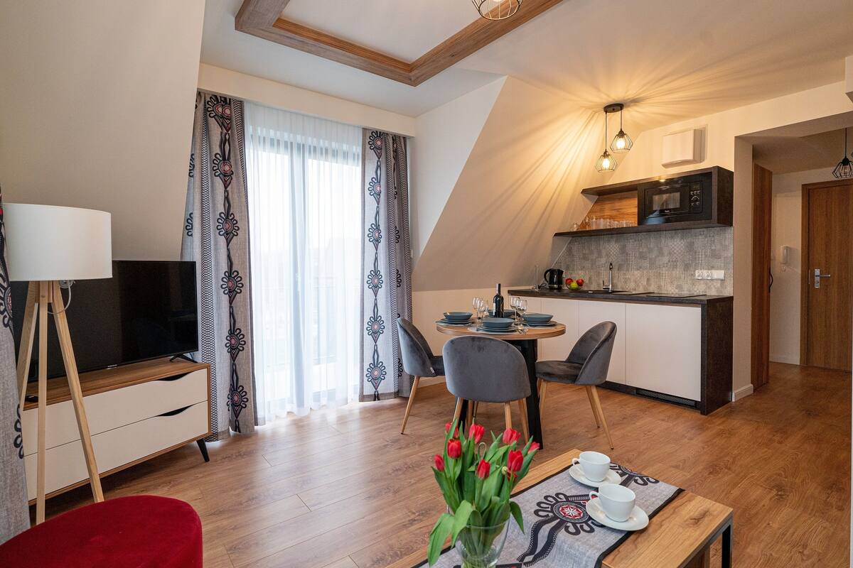 Uma sala de estar moderna com televisão, um espaço de jantar com mesa posta para dois e uma cozinha com micro-ondas.
