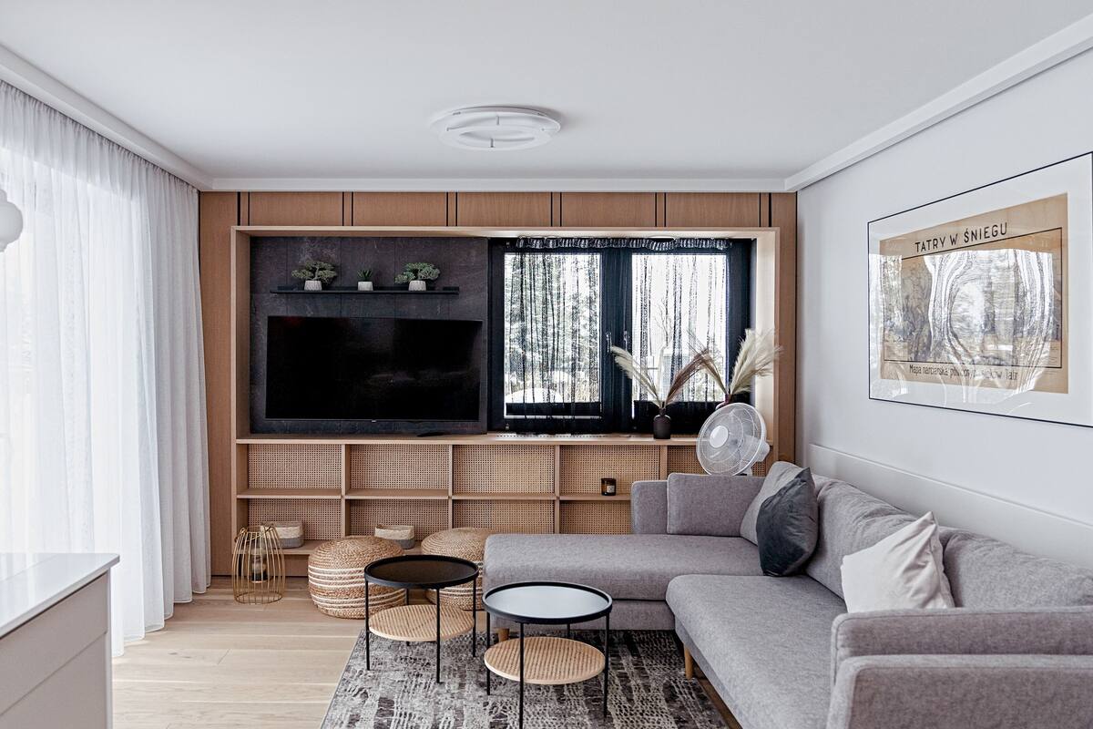 Uma sala de estar moderna com um sofá cinzento, uma TV de tela plana fixada em um painel de madeira, uma janela grande com cortinas leves e uma obra de arte emoldurada na parede.