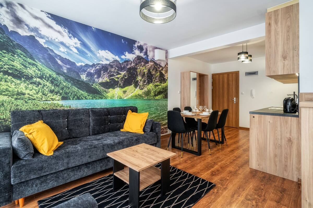 Uma sala de estar moderna com um sofá cinza, uma mesa de centro de madeira e uma área de jantar com mesa e cadeiras de madeira. Um mural grande com paisagem de montanha está na parede.