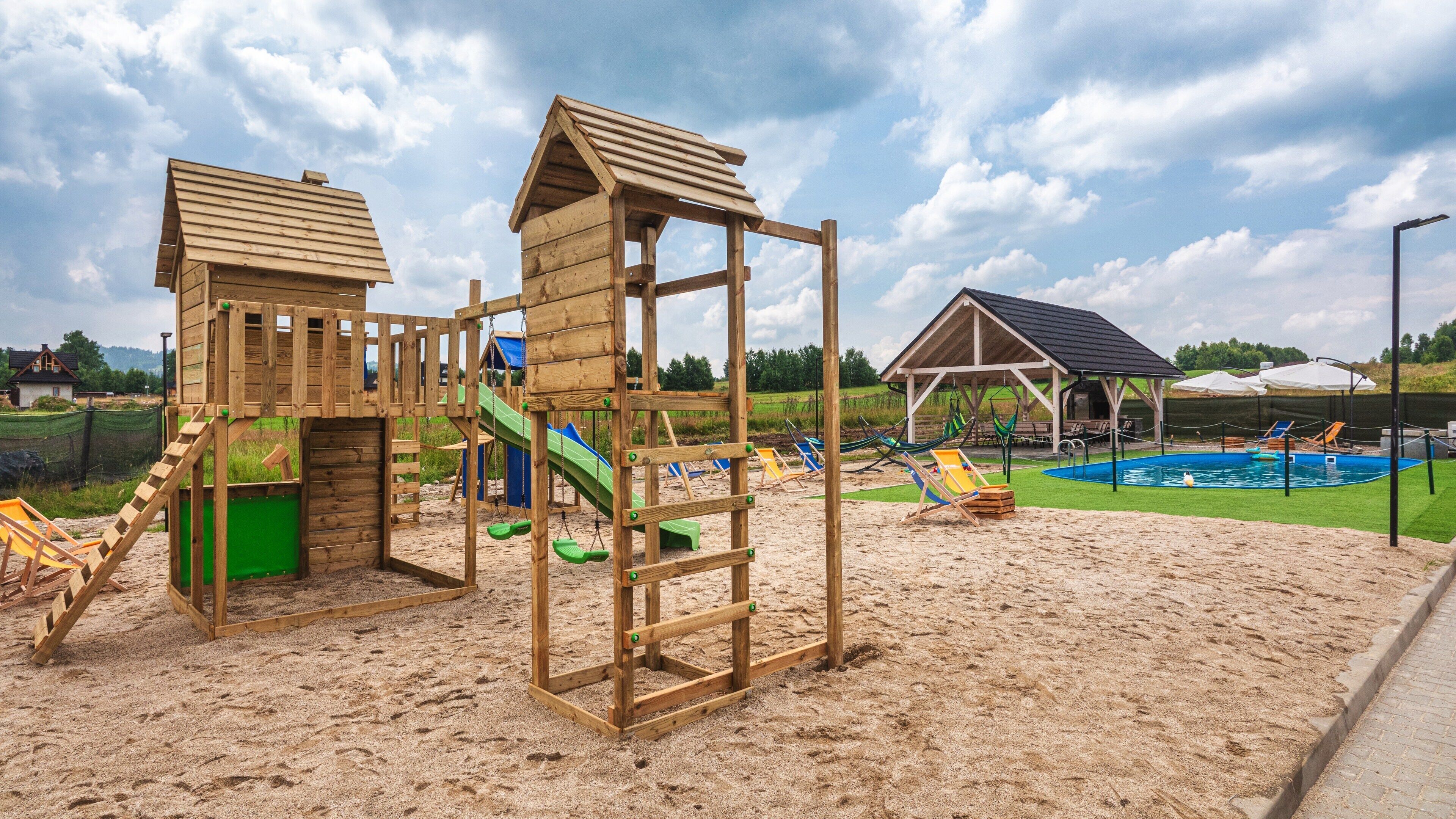 A wooden play structure with slides and climbing elements, a small pool with lounge chairs, and a covered seating area.