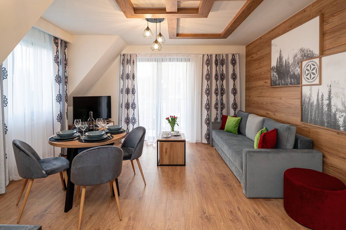 Uma sala de estar moderna com área de jantar, apresentando teto de madeira, sofá cinzento e um banquinho vermelho.