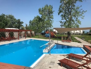 A swimming pool with a wooden deck, lounge chairs, and a playground in the background. - LB - Silver Seaside Apartment (Siófok)
