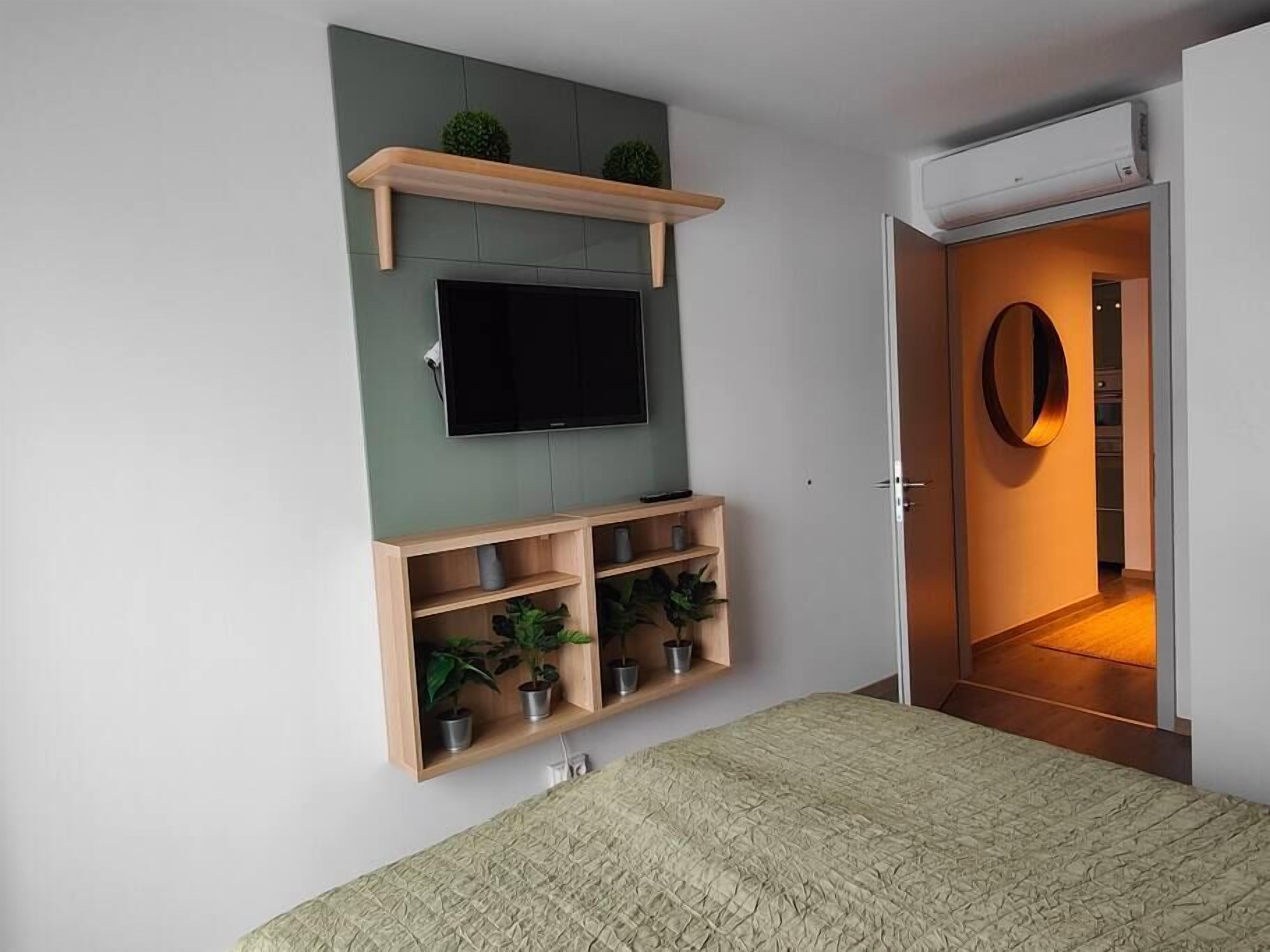Een slaapkamer met een bed, een wandtelevisie, een plank met planten en een deur naar een andere kamer.