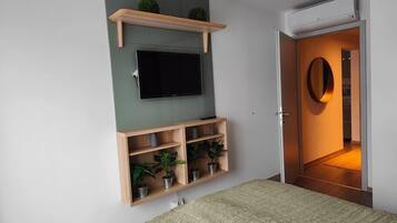 Un dormitori amb un llit, una televisió penjada, una estant amb plantes i una porta que dona a una altra habitació.