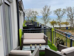 A balcony with outdoor furniture, a glass table, and a view of a body of water and trees. - LB - Ezüst Panoráma Lovely 1 bedroom condo on the beach (Siófok)
