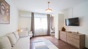 A modern living room with a sofa, a TV, a wooden cabinet, and a large window with curtains. - LB - Phoenix Apartman Siófok (Siófok)