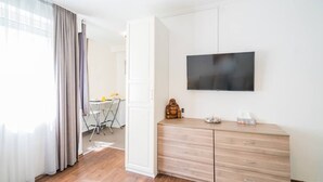 A hotel room with a wooden dresser, a flat-screen TV mounted on the wall, and a dining area visible through an open door. - LB - Phoenix Apartman Siófok (Siófok)