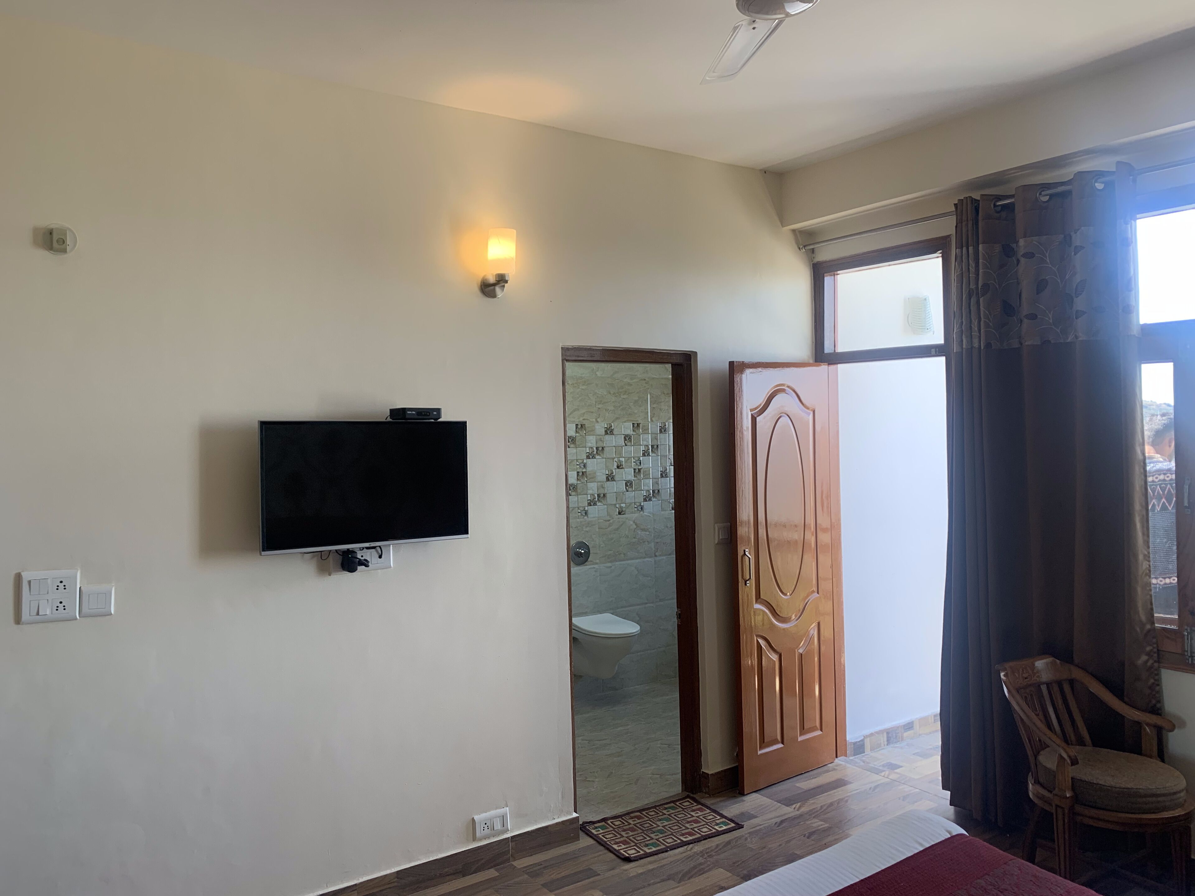 A room with a flat-screen TV mounted on the wall, a wooden door, a chair, and a window with curtains.