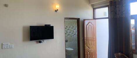 A room with a flat-screen TV mounted on the wall, a wooden door, a chair, and a window with curtains.