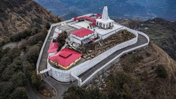 An aerial view of a large, fortified structure with a central tower, surrounded by buildings with red roofs, set against a mountainous backdrop.