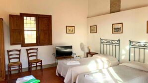 Quarto com duas camas, televisão sobre suporte, cadeiras de madeira e uma mesa com uma pasta verde.