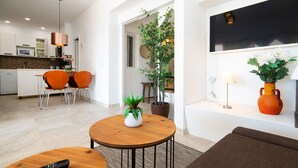 Un salon moderne avec une table basse en bois, un canapé et un téléviseur fixé au mur. On y trouve également un coin cuisine avec des meubles blancs et une table à manger avec des chaises.