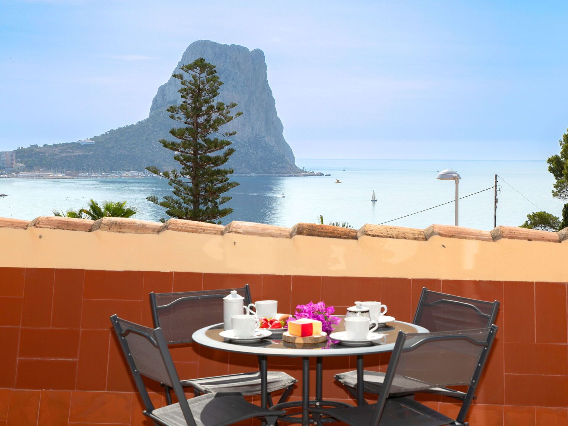 A table with four chairs and a table setting including cups, a teapot, and a plate with food and flowers. A mountain is visible in the background.