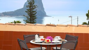 Une table avec quatre chaises et une mise en place comprenant des tasses, une théière et une assiette avec de la nourriture et des fleurs. On aperçoit une montagne à l’arrière-plan.