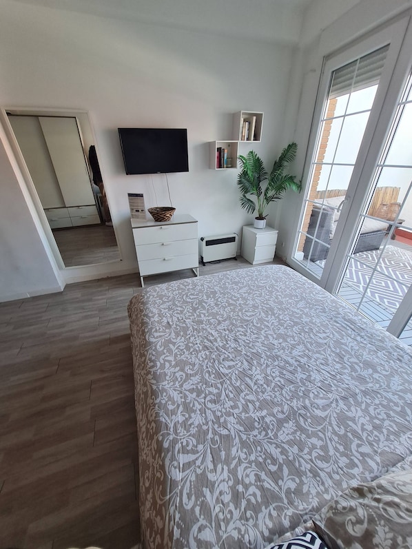 A bedroom with a large bed, a flat-screen TV mounted on the wall, a potted plant, and a sliding glass door leading to an outdoor balcony.