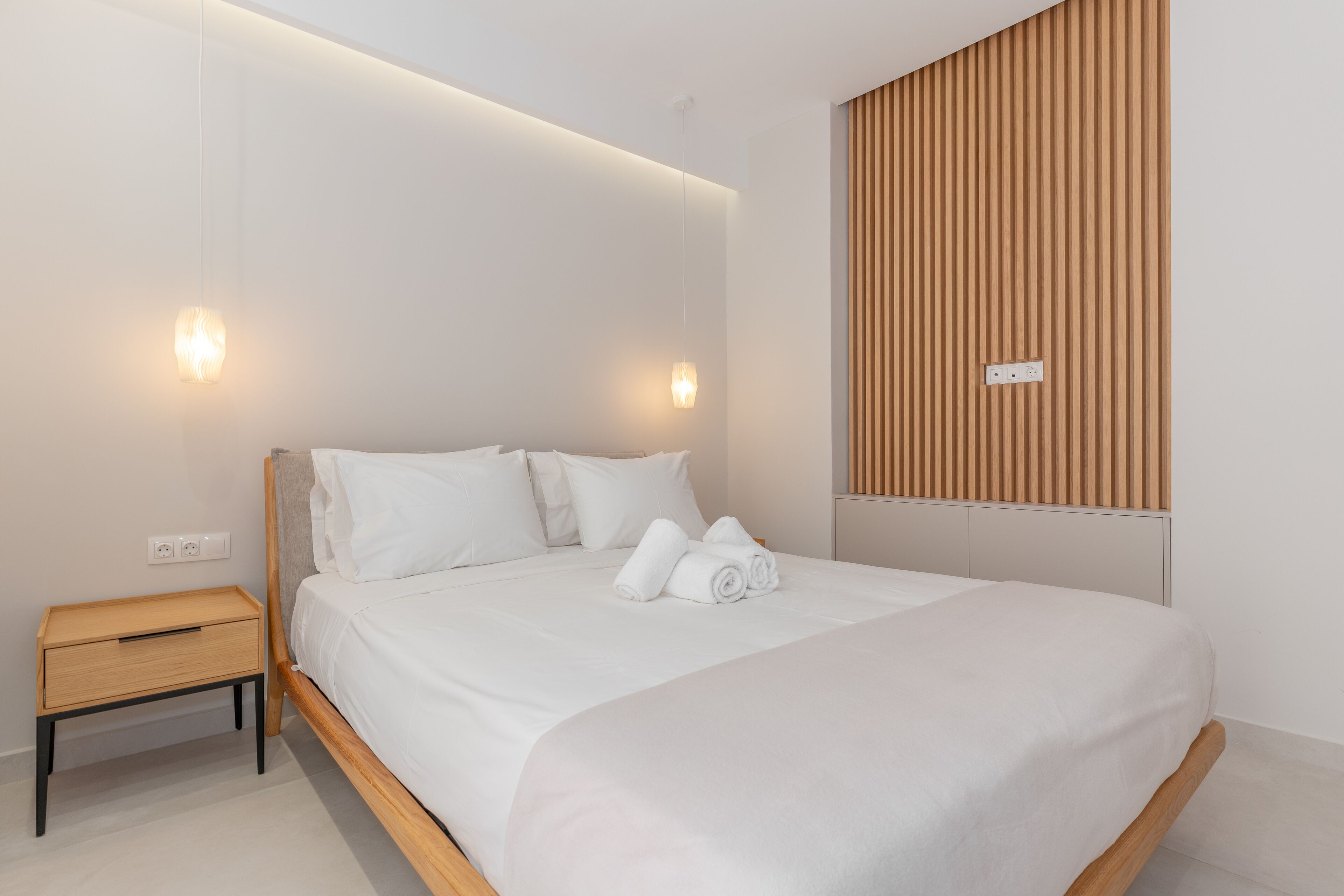 Un dormitorio moderno con cabecera de cama de madera, ropa de cama blanca y dos mesitas de noche.