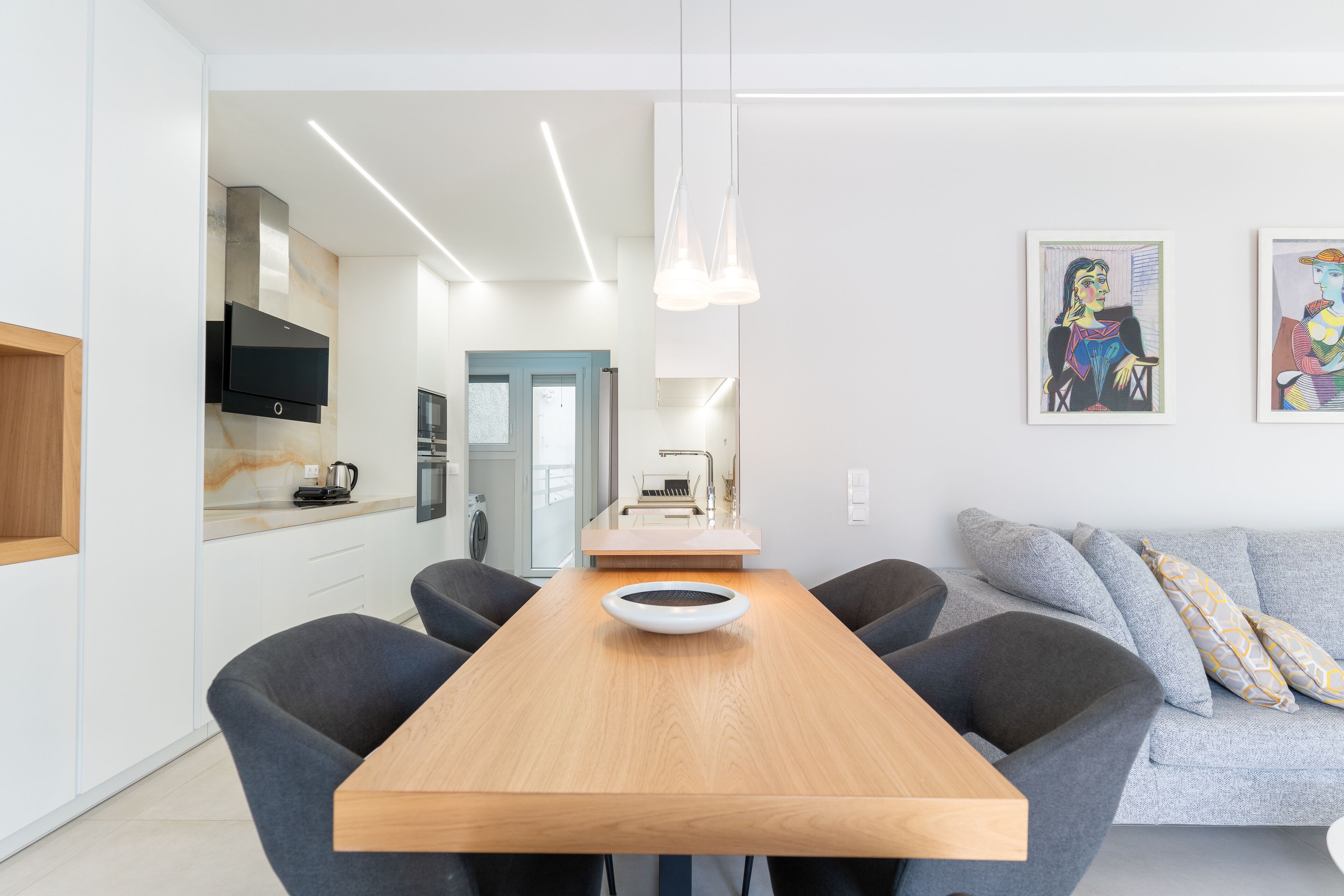 A modern kitchen with a wooden table, grey chairs, and a white bowl. There is a grey sofa with patterned cushions in the living area.
