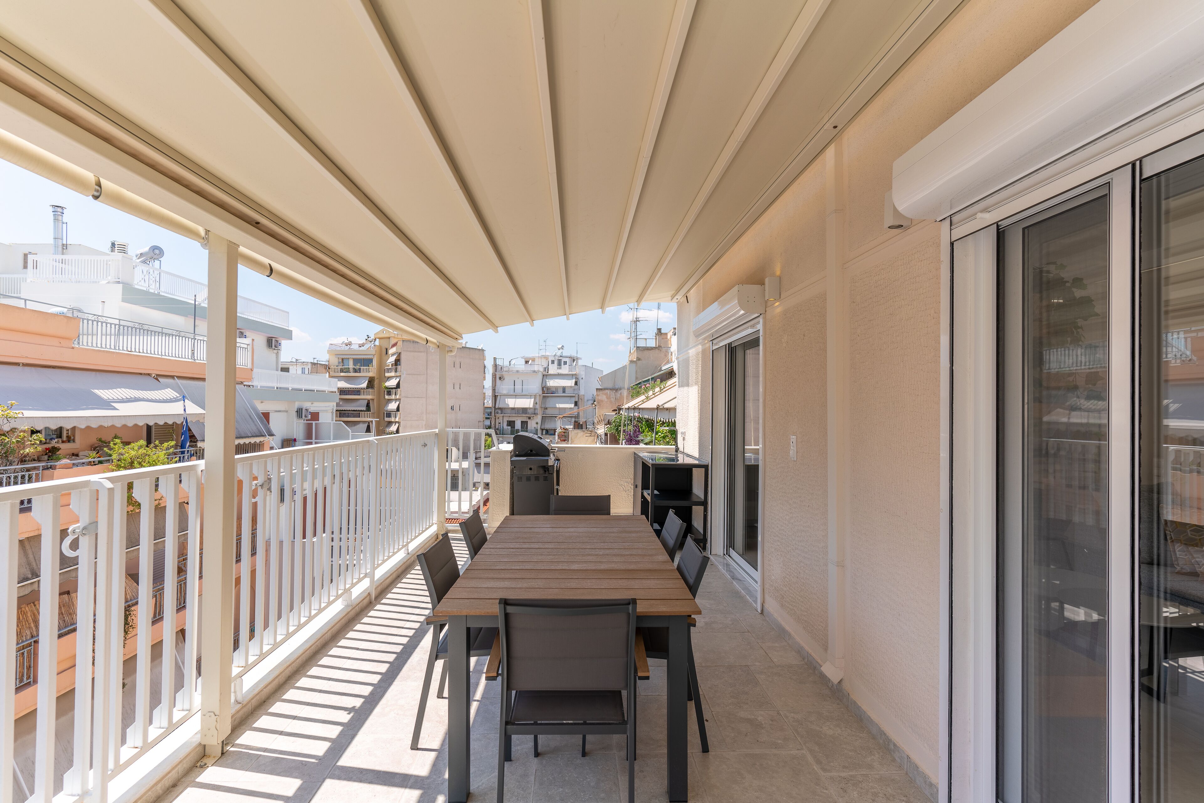 A balcony with a wooden table and chairs, a grill, and a view of neighboring buildings.