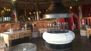 A rustic interior with wooden furniture, a large central stove, and a thatched roof.