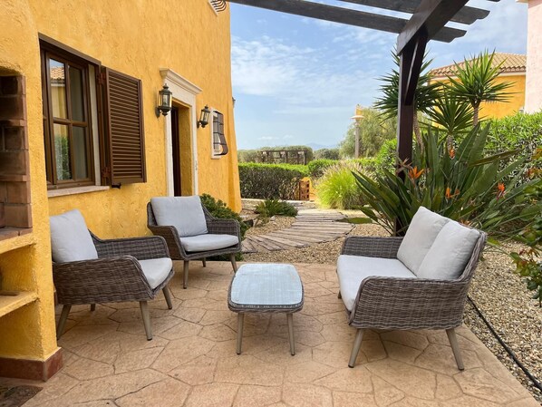 An outdoor patio with wicker furniture, a stone floor, and a yellow building with windows and doors.