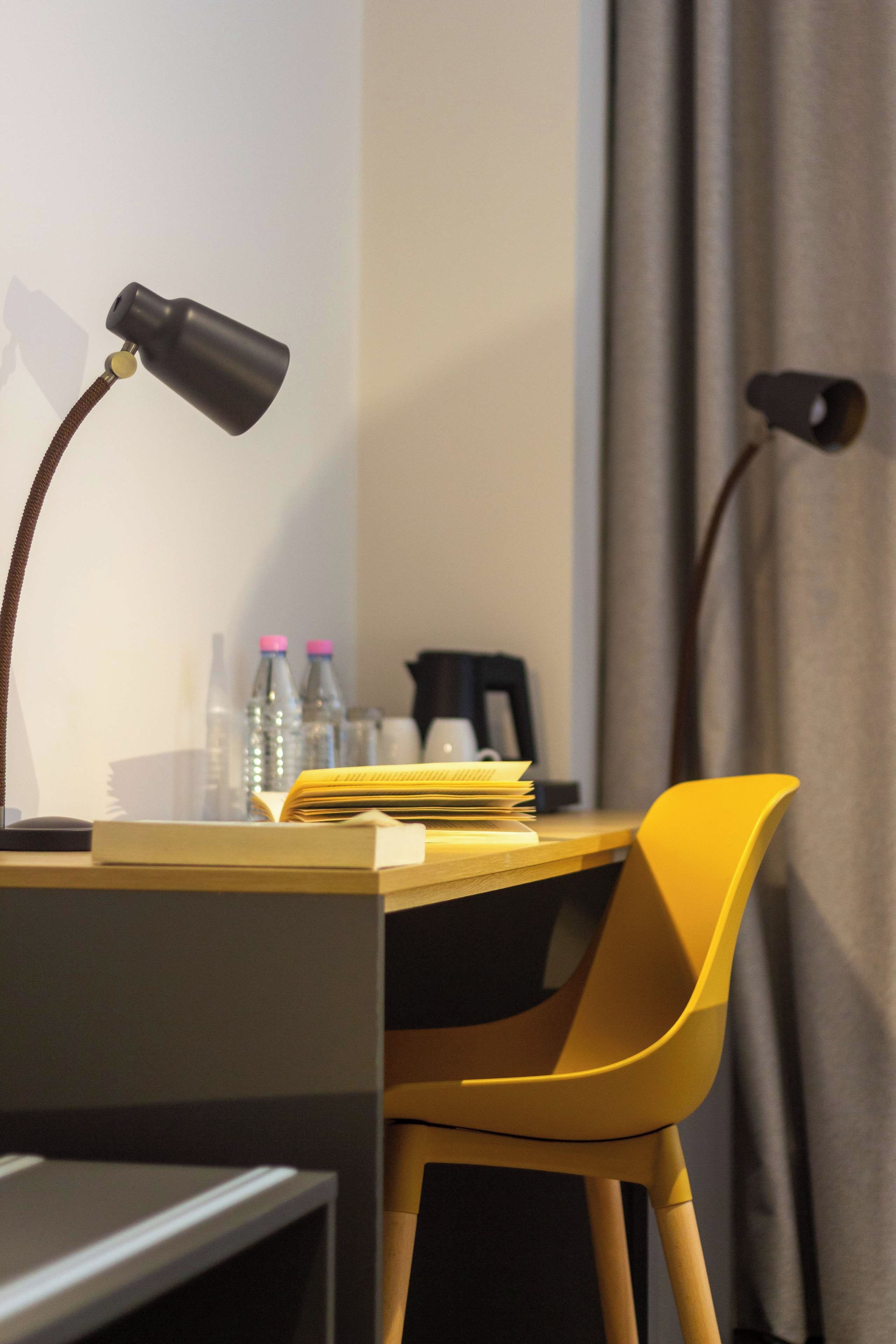 A desk with a lamp, a chair, a stack of papers, and two bottles on it.