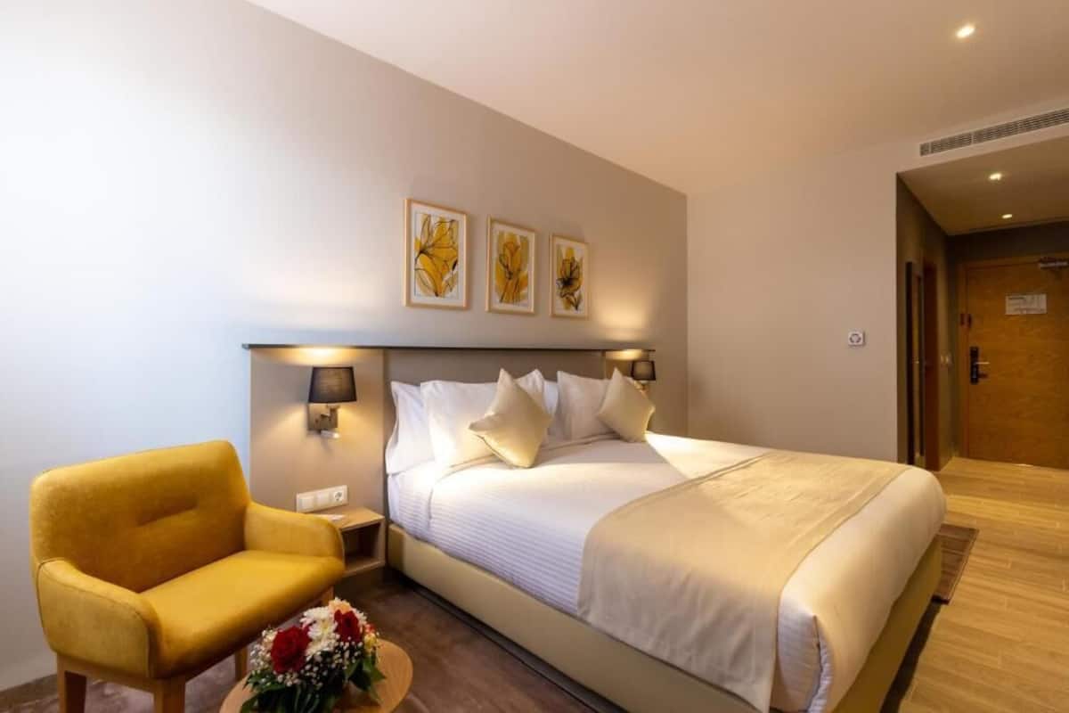 A hotel room with a bed, a yellow armchair, a small table with a vase of flowers, and three framed pictures on the wall.