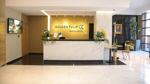 En receptionsdisk på Golden Tulip Opera Hotel med en marmorbordplade, indrammede kunstværker og potteplanter.