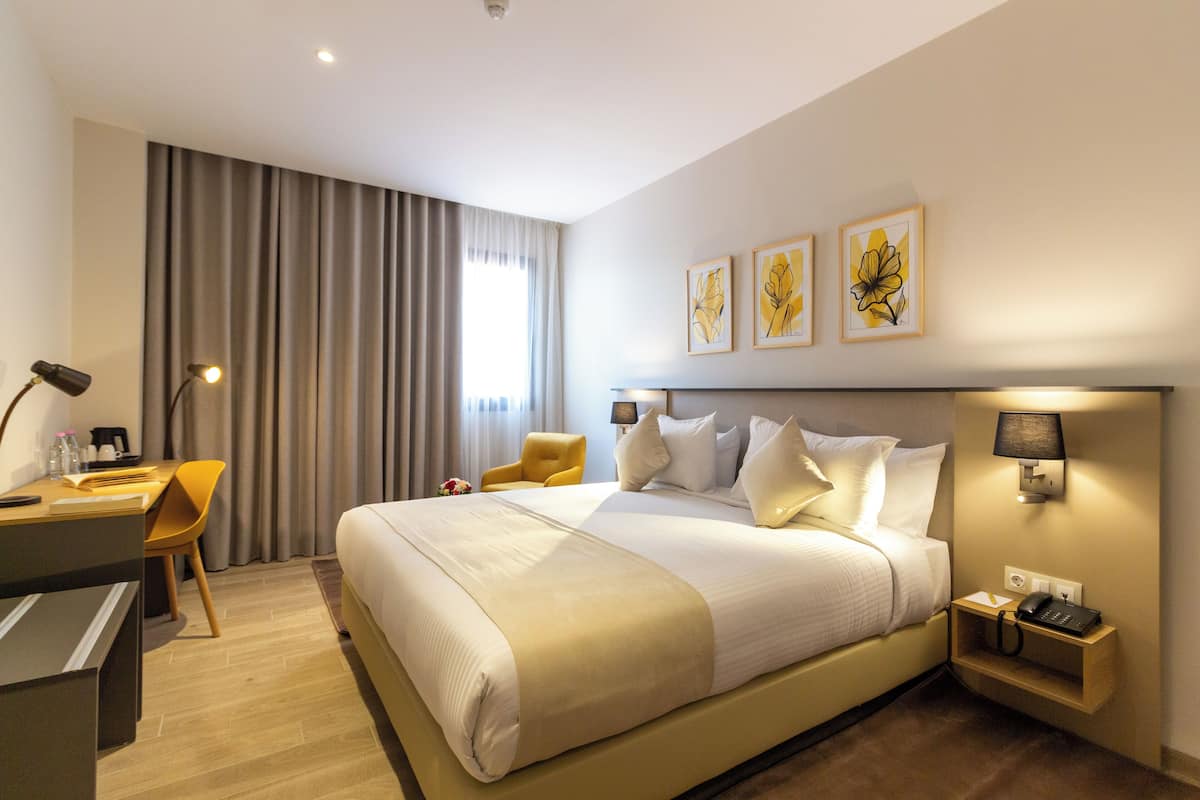 A hotel room with a large bed, a desk with a chair, a nightstand with a lamp, and artwork on the wall.