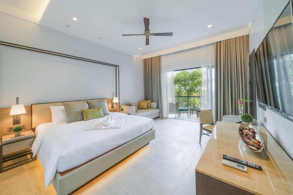 A modern hotel room with a large bed, a desk with a remote control, and a view of greenery outside.