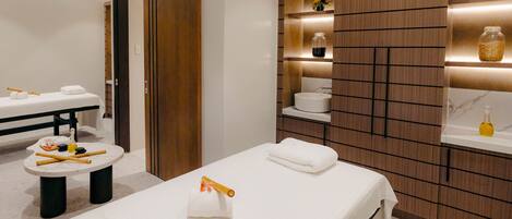 A modern spa room with a massage table, a small round table with towels and massage tools, and wooden paneling with shelves holding jars and a small plant.