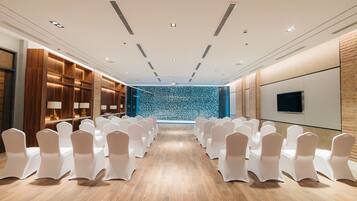Un salón de conferencias con filas de sillas blancas, suelo de madera, pared de piedra y un televisor montado.